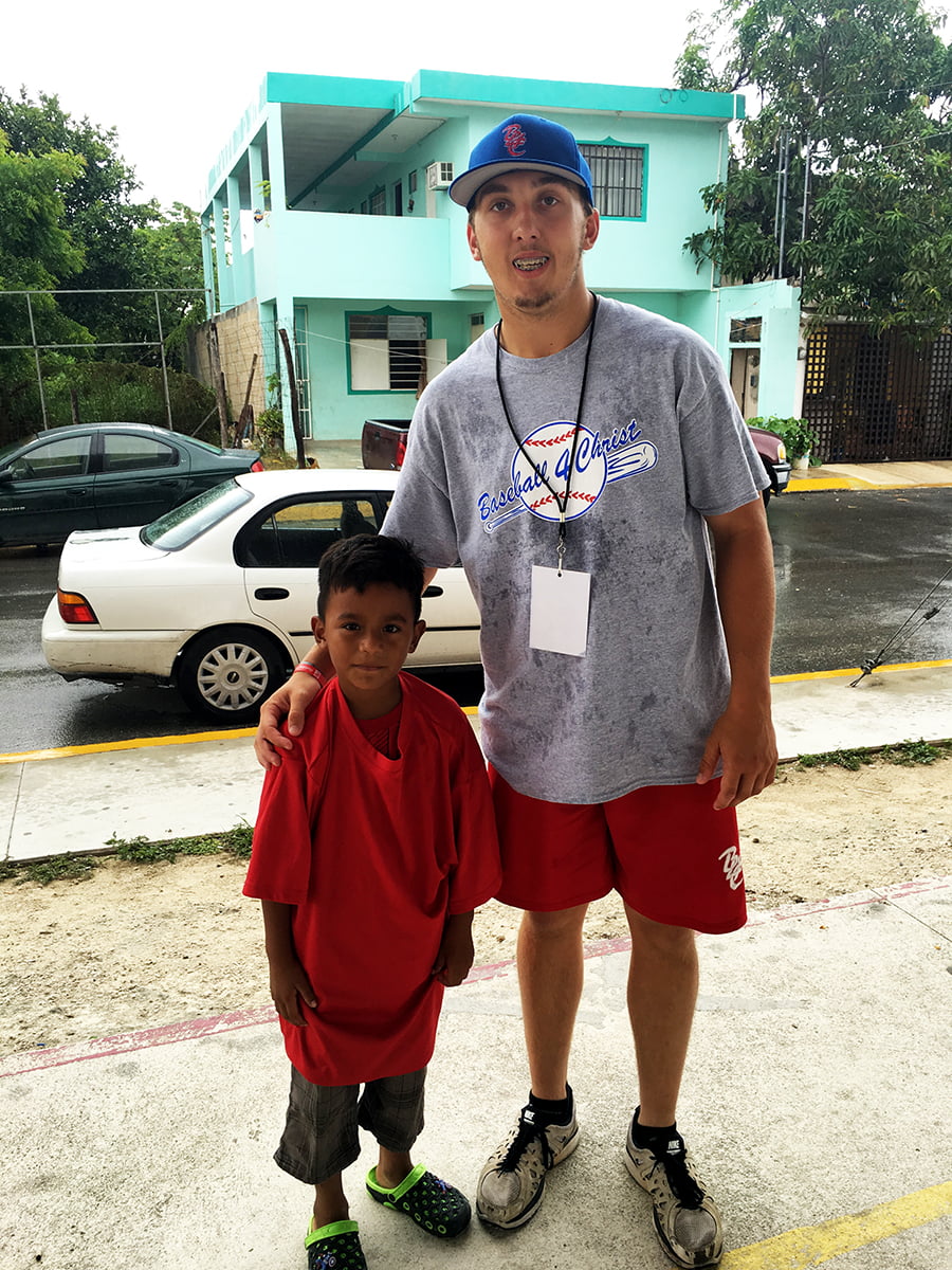 A baseball 4 christ team player standing next to a parked child in Mexico.